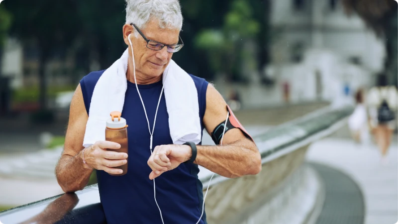 Um homem idoso, com o corpo atlético, descansa após uma corrida. Ele segura uma garrafa de energético, uma toalha pendurada no pescoço com um fone de ouvido, enquanto reflete sobre como envelhecer bem."