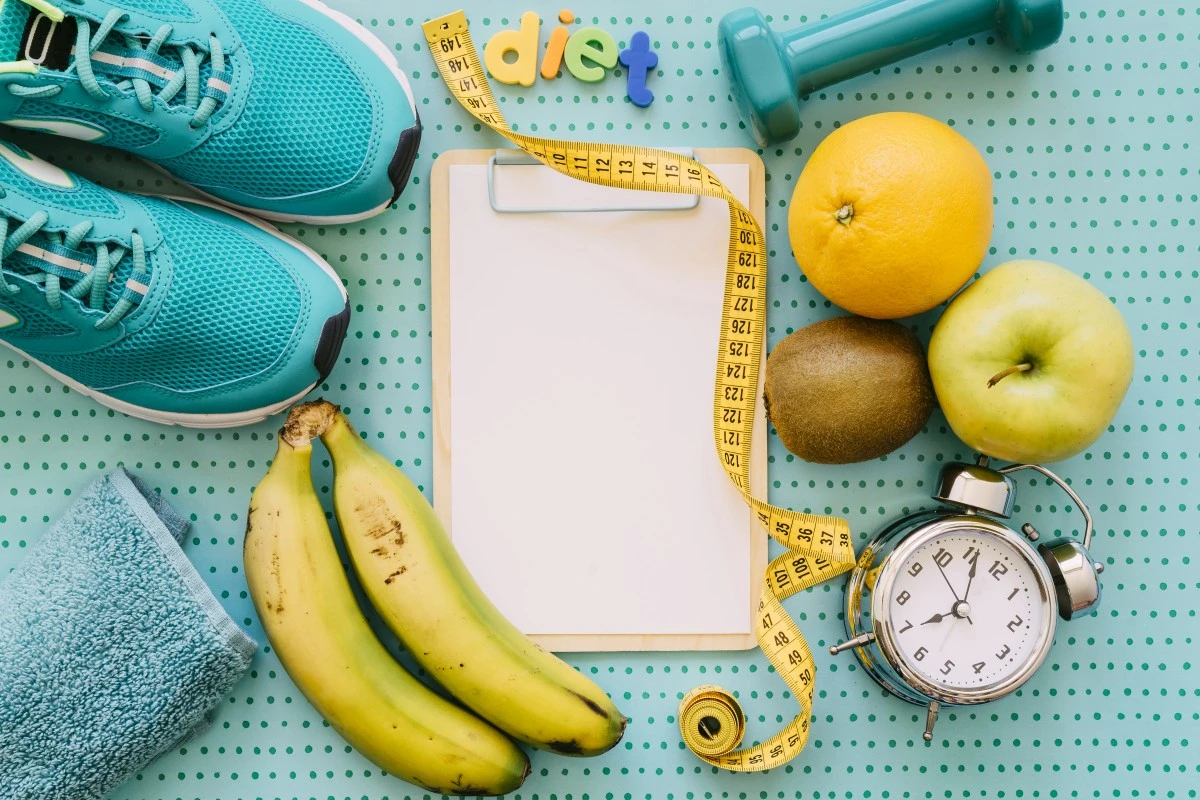 Um fundo escrito verde escrito diet, um par de tênis verde, uma toalha de rosto verde, duas bananas, uma fita métrica, um peso de academia, uma laranja, uma maçã, um kiwui e um dispertador analógico. Símbolos de uma jornada fitness rumo à perda de peso com atividade física.