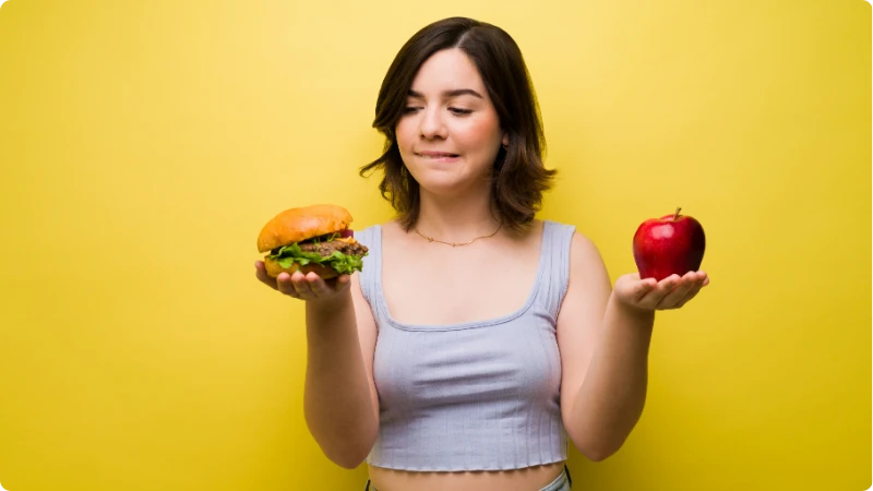 Decisões alimentares: uma mulher segura uma maçã em uma mão e um sanduíche na outra, refletindo sobre a importância da alimentação balanceada na prevenção de doenças.