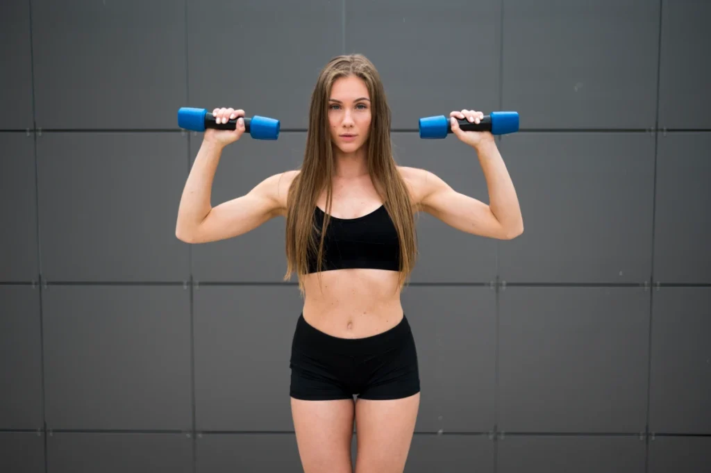 Fitness mulher com roupas esportivas segurando halteres e se preparando para fazer exercícios de desenvolvimento dos ombros. 10 Exercícios para Iniciantes na Academia e em Casa.
