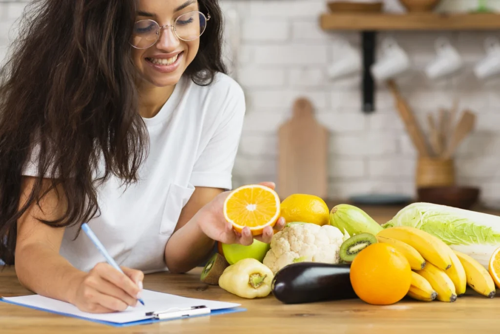 Dieta Low Carb: o que é, como fazer, benefícios e cardápio. Vários alimentos saudáveis sobre a mesa e uma moça jovem sorridente, fazendo anotações do seu planejamento alimentar.