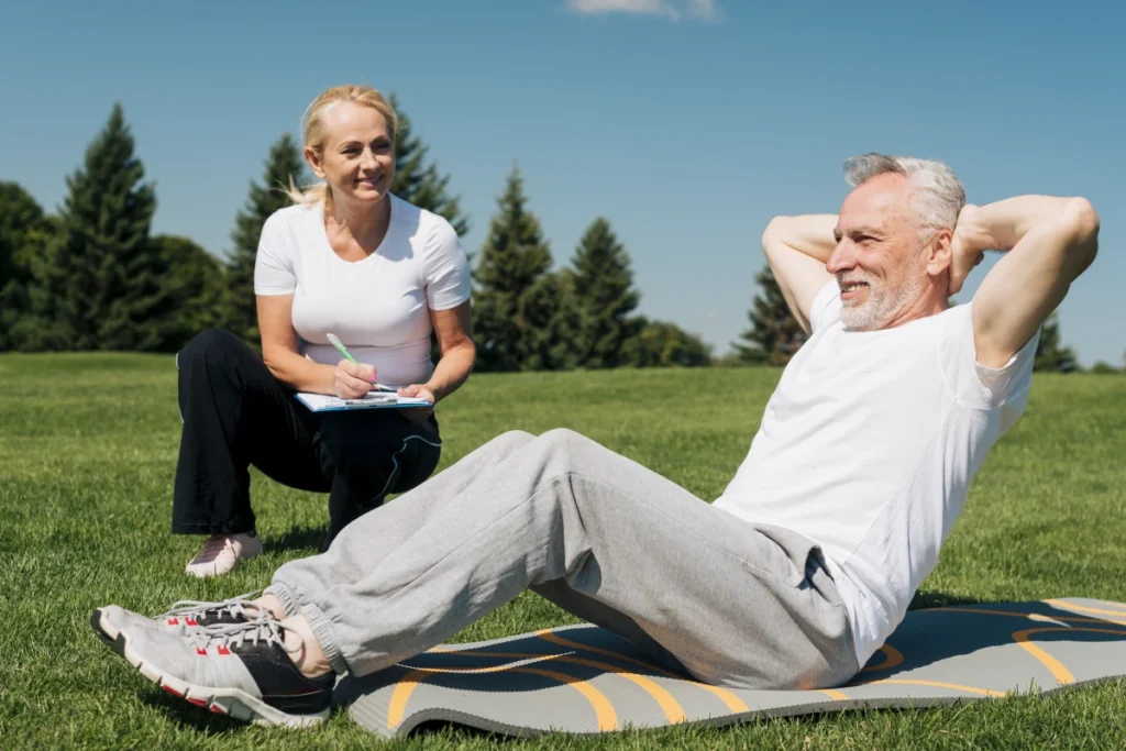Envelhecimento Saudável: O que é, Fases, Benefícios e Dicas. Dois idosos, um homem e uma mulher em um parque. Dois idosos, um homem e uma mulher, em um parque. O homem faz exercício abdominal, enquanto a mulher o observa e faz anotações em uma prancheta.
