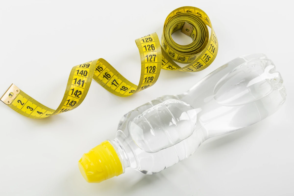 Beber Água Emagrece? Descubra Como Acelerar a Perda de Peso. Garrafa de água vista do alto com uma fita métrica amarela.