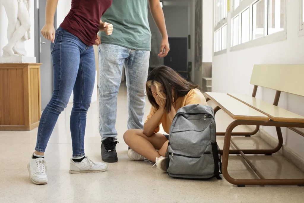 Ansiedade: O que é, causas, sintomas e tratamento. Garota sentada no chão com as mãos no rosto sendo intimidada na escola por dois colegas.