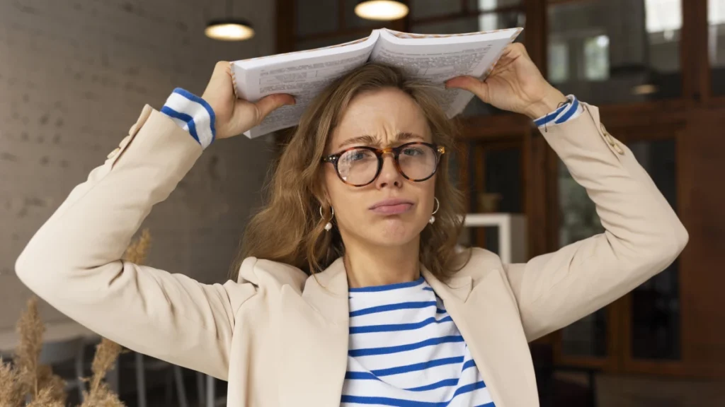 Cansaço Mental: Como Identificar, Entender e Combater a Exaustão da Mente. Mulher jovem com um livro na cabeça demonstrando irritação e cansaço no trabalho.