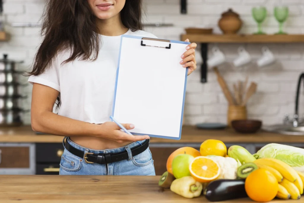 Cardápio para Emagrecer: Plano de 5 Dias Completo. Mulher morena close-up segurando uma prancheta em uma cozinha e uma bancada com várias frutas.
