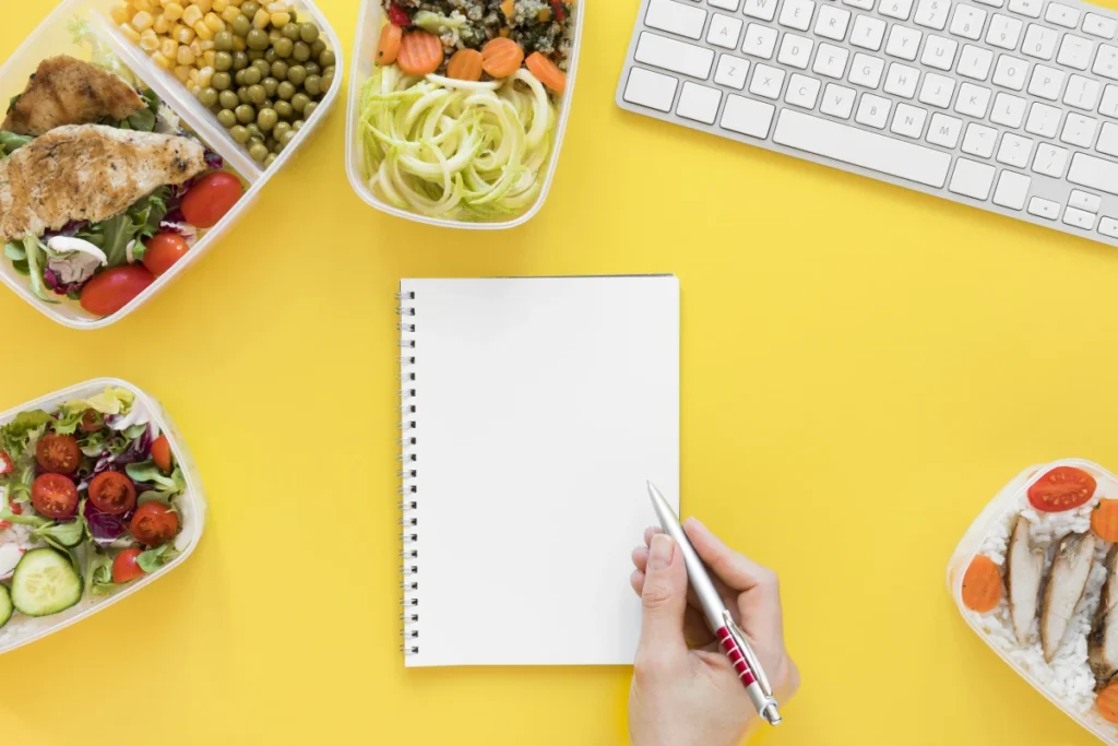 Diário Alimentar: O Que é, Como Funciona e Como Fazer. Várias porções de alimentos saudáveis, imagem de uma mão fazendo anotações em um caderno e um teclado de computador sobre a mesa amarela.