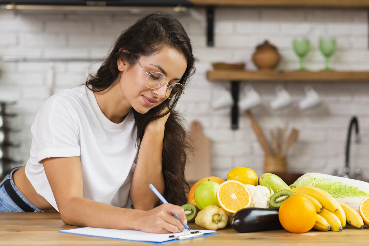 Diário Alimentar: O Que é, Como Funciona e Como Fazer. Uma mulher jovem de cabelo negros longos, fazendo anotações em uma prancheta na bancada da cozinha com vários alimentos saudáveis próximos.
