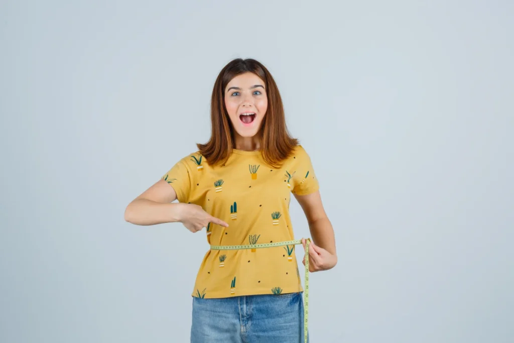 Dieta Mediterrânea: O Que é, Benefícios e Como Fazer. Moça jovem, alegre, comemorando a perda de peso ao medir a cintura com uma fita métrica.