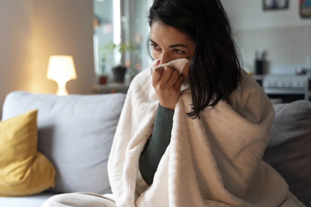 Doenças Respiratórias no Inverno Como Prevenir. Uma mulher sentada no sofá da sala, com coberta e usando um lenço para limpar o nariz devido ao resfriado.