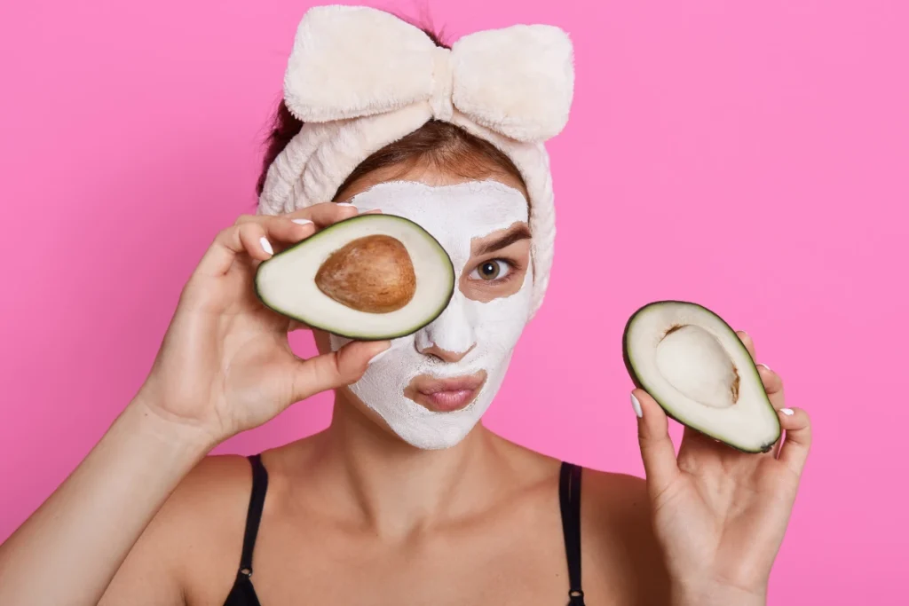 Envelhecimento Precoce: Causas, Sintomas e Prevenção. Mulher bonita spa com mascara facial no rosto e segurando metades de abacate nas mãos olhando para câmera fazendo procedimentos de cosmetologia em casa usando faixa de cabelo com laco.