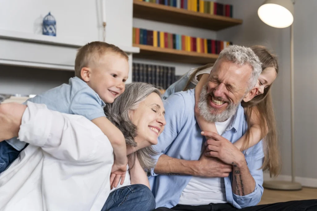 Idoso Feliz: Como Ter Alegria e Bem-Estar com 8 Dicas. Um casal de idosos felizes com os netos pequenos na sala de casa.