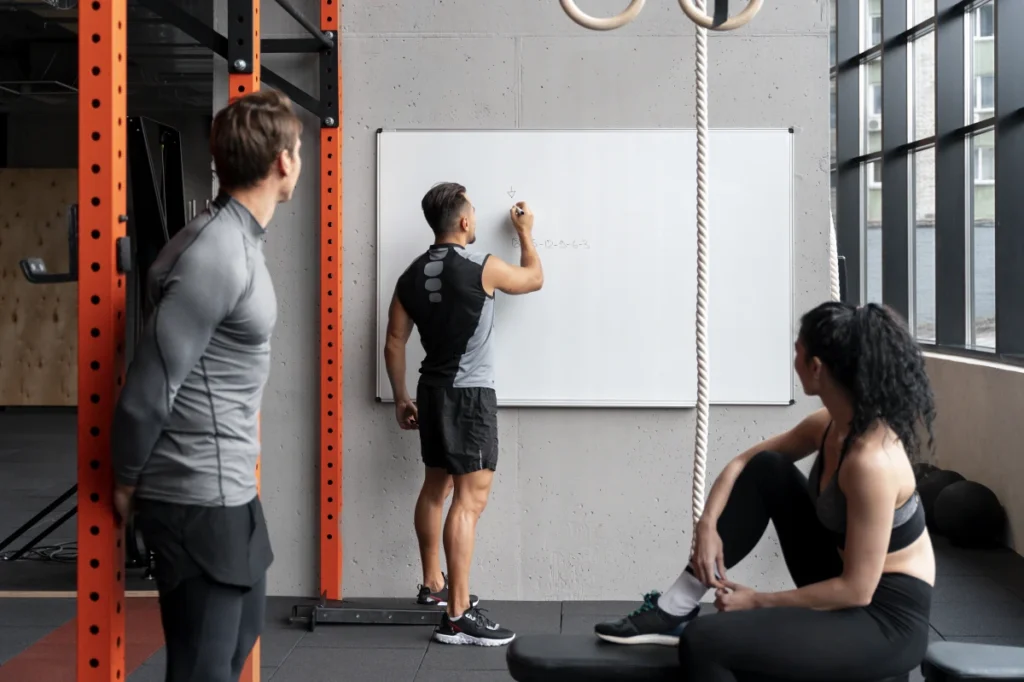 Treino ABC: O Que É, Como Fazer, Benefícios e Cronogramas. Pessoas na academia conversando e fazendo planos de exercícios em um quadro na academia.