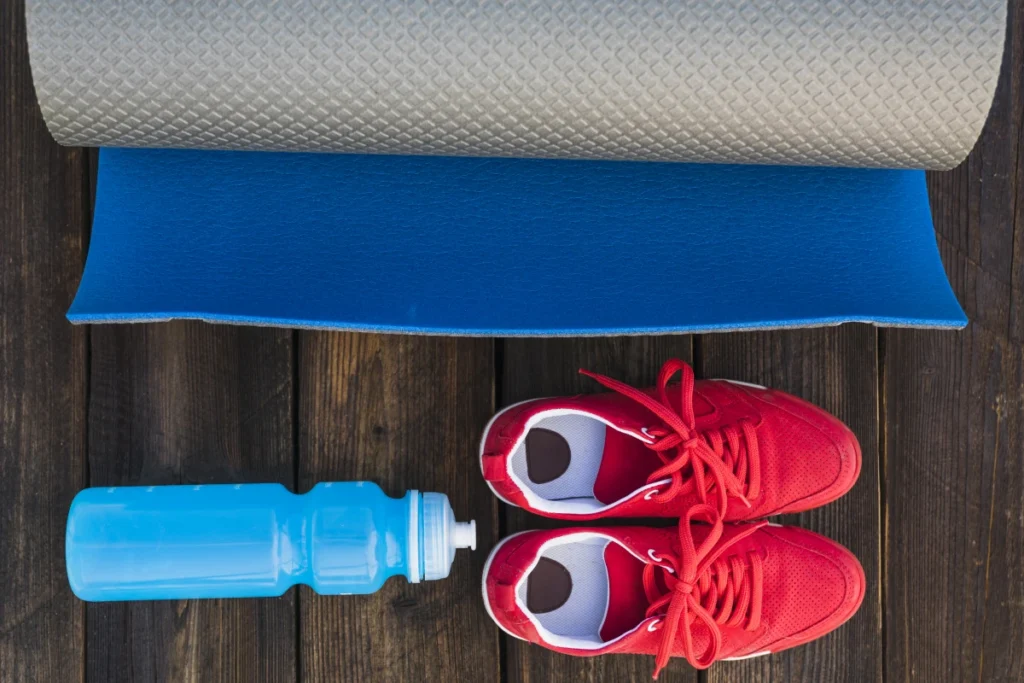 Treino Tabata: 4 Minutos Para Transformar Seu Corpo. Rolo de tapete para exercícios, com garrafa de água e par de tênis esporte sobre um piso de madeira.