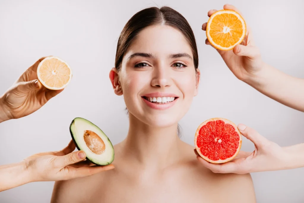 Vitaminas: O que são, para que servem, tipos e classificação. Pele radiante linda e saudável de mulher sem maquiagem retrato de menina sorrindo contra parede de frutas.