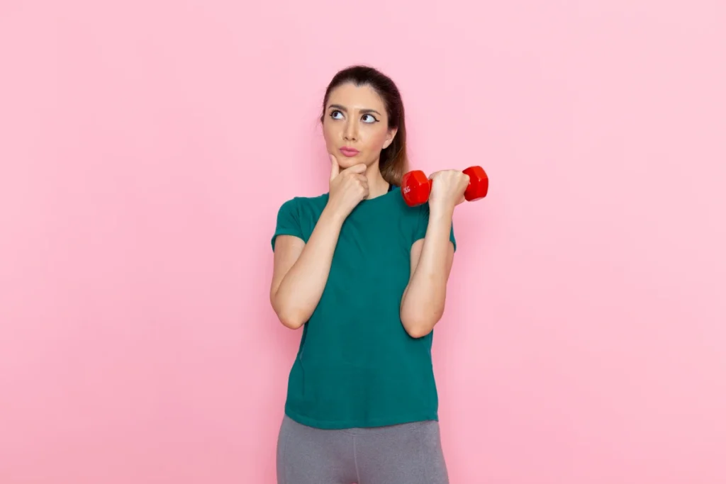 Zumba: O Que É, Benefícios e Como Começar. Mulher jovem segurando halteres na parede rosa demonstrando estar com dúvida.