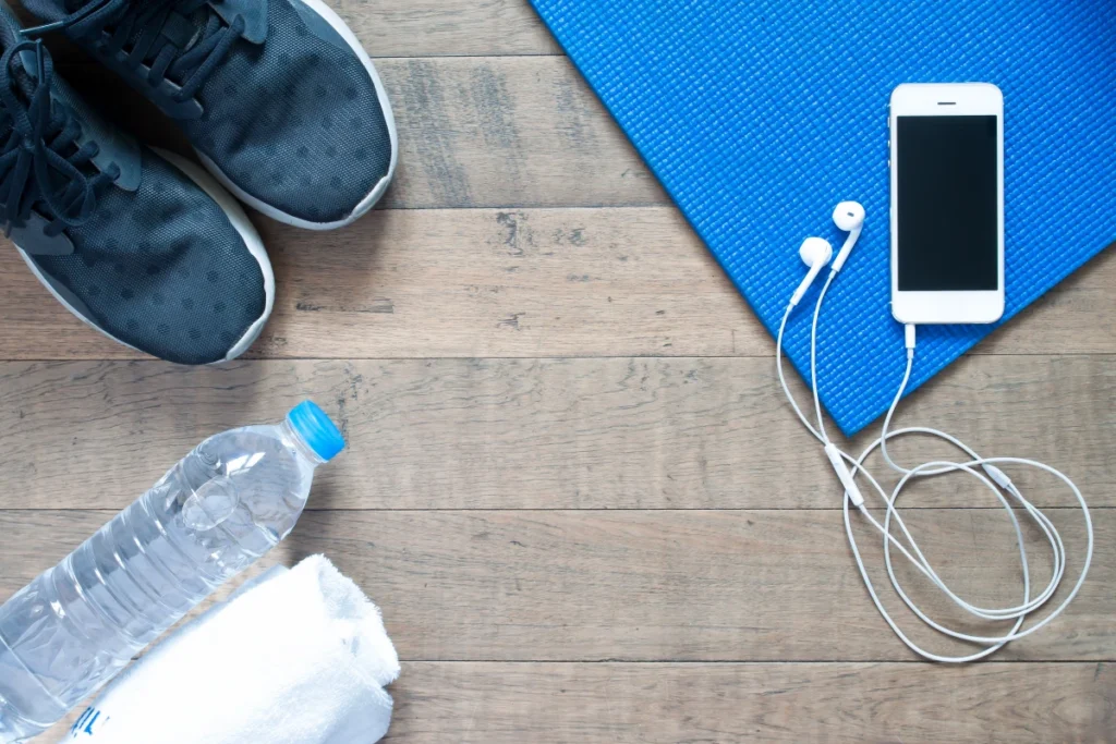 Treino Intervalado: o que é, tipos e como fazer. Vista aérea de um smartphone com fone de ouvido em um tapete azul para exercícios no solo, um tênis, uma garrafa de água e uma toalha branca.