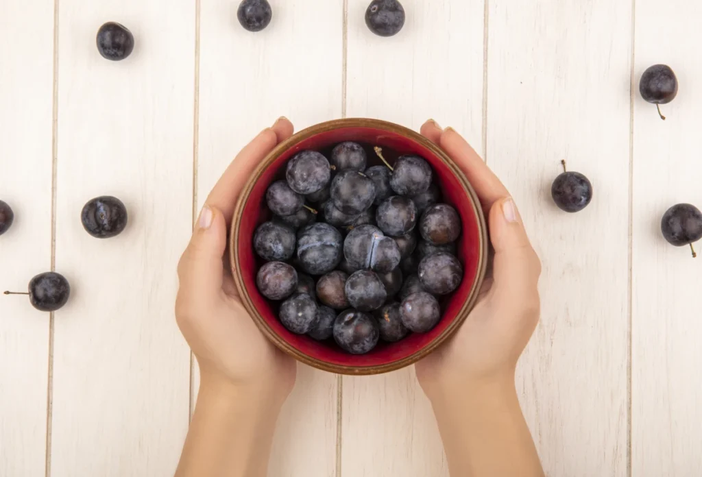 A Fruta Roxa que os Médicos Recomendam para Quem Sofre com Intestino Preso 1. Vista superior de mãos femininas segurando uma tigela com várias ameixas escuras dentro.